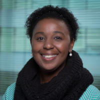 A foto mostra o rosto de uma mulher sorridente sobre um fundo verde-água. Ela é negra, tem cabelos curtos e escuros e olhos também escuros, e veste um cachecol preto sobre uma camisa verde-água.